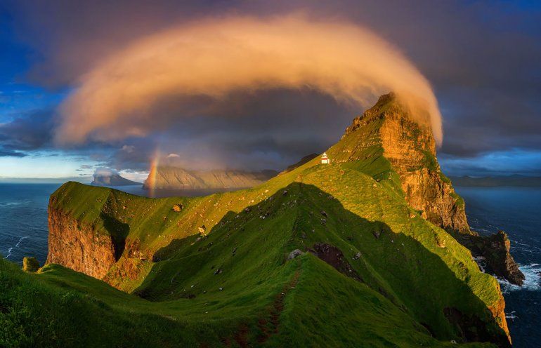 La puesta del sol ilumina un faro y arco iris en las Islas Feroe.